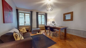 A living room with a sofa, a dining table with chairs, a chandelier, and large windows with curtains. - Gite de Paul - centre ville Colmar (Colmar)