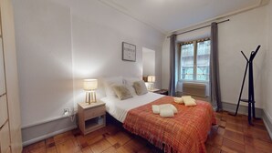 A hotel room with a bed, two bedside tables, a standing lamp, a window with curtains, and a radiator. - Gite de Paul - centre ville Colmar (Colmar)