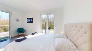 A bedroom with a large bed, a chair, a picture on the wall, and a view of the outdoors.