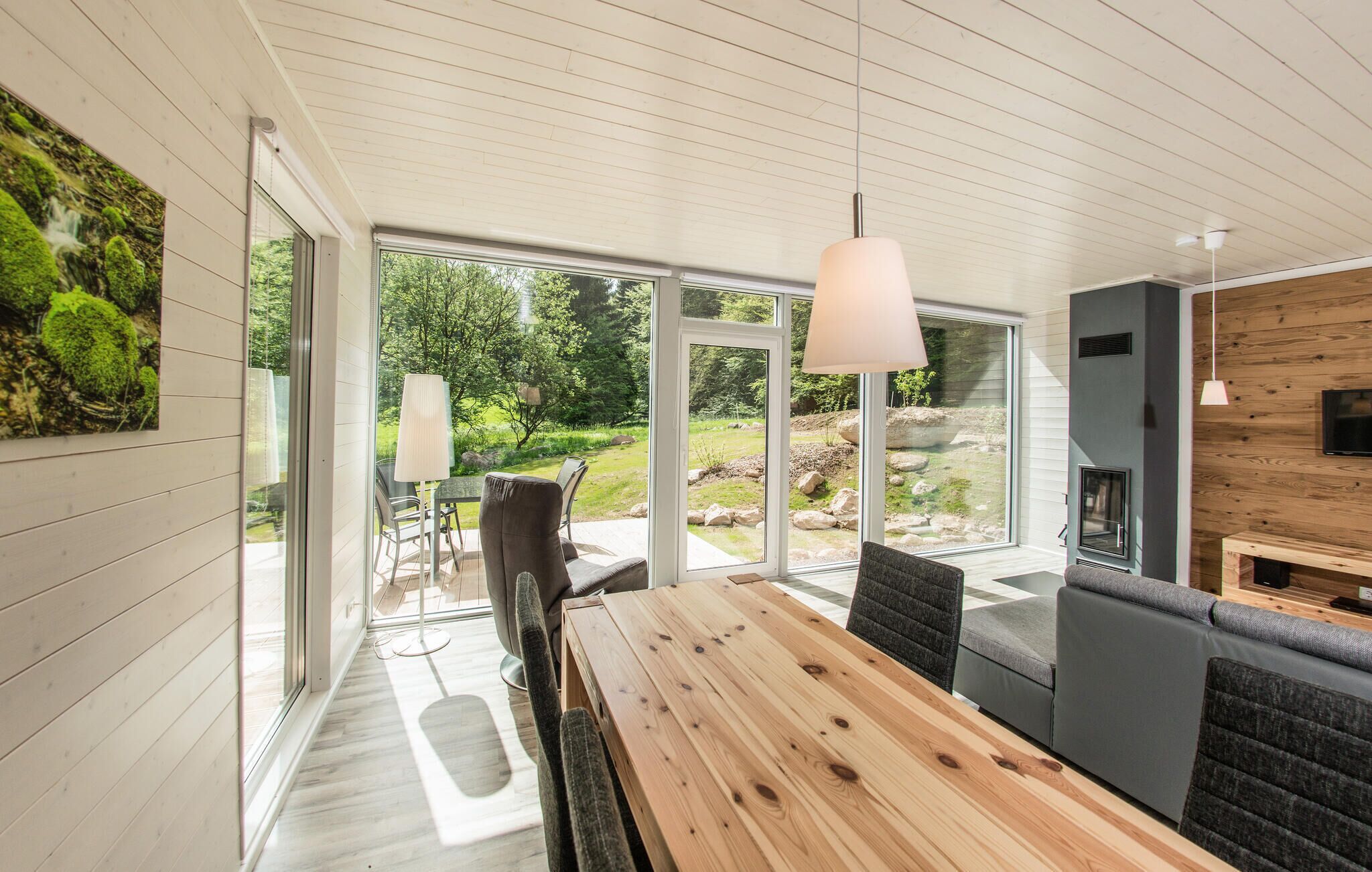 Ein modernes Interieur mit einem Holzesstisch, Stühlen und einer Couch. Große Fenster bieten Blick auf einen Garten.