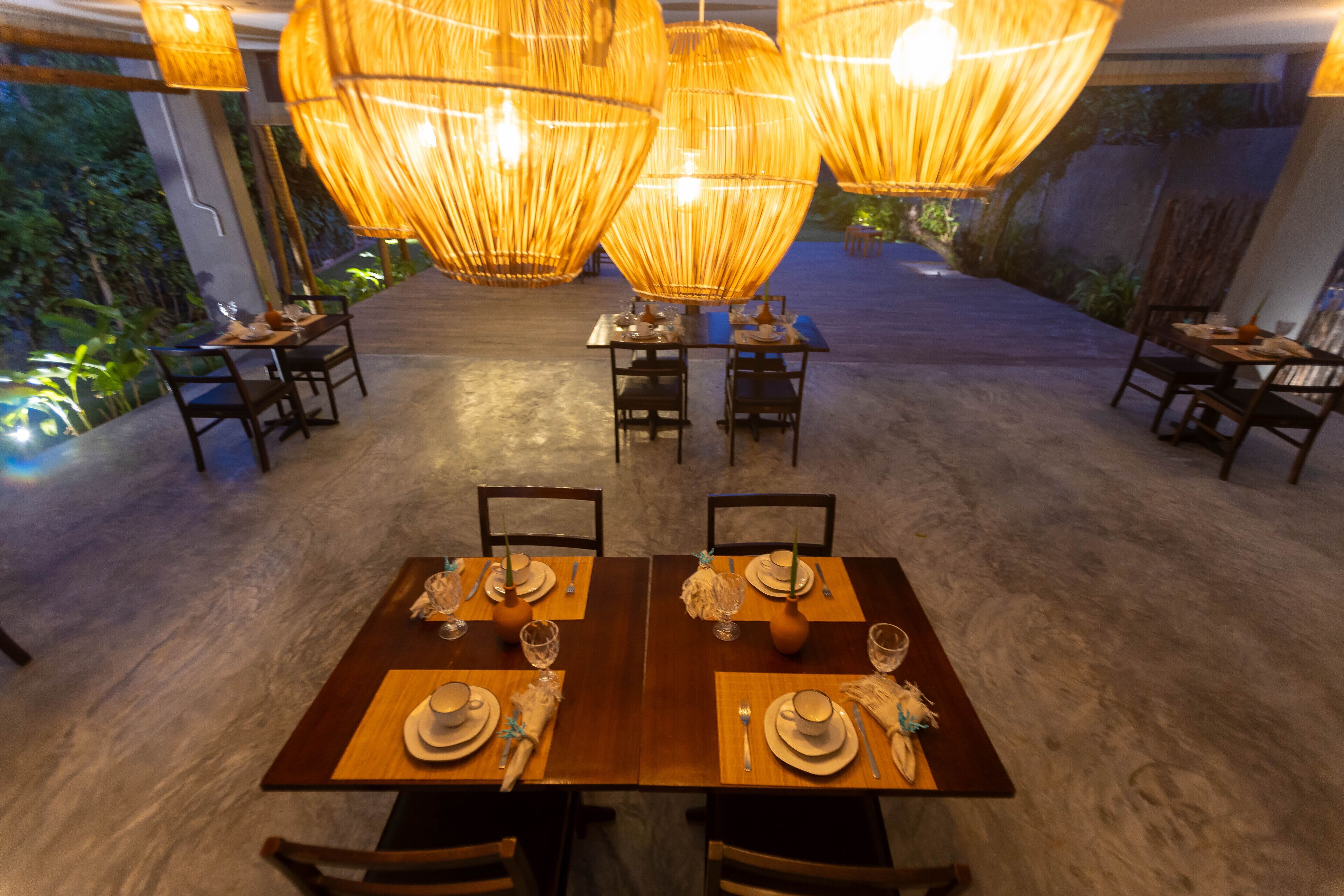 A dining area with tables set for two, featuring woven light fixtures and a view of greenery outside.