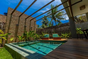Piscina com deck de madeira e espreguiçadeiras, cercada por plantas tropicais e um muro de pedra.