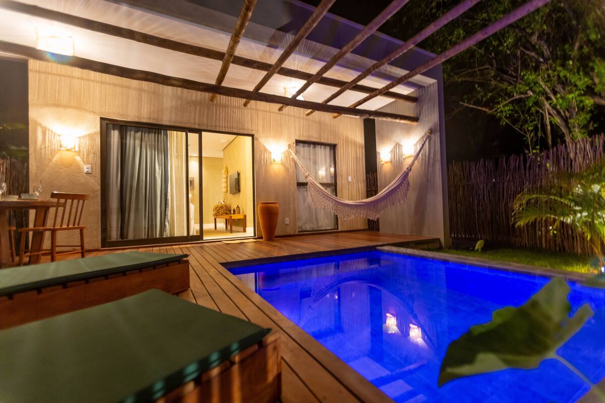 A pool area with wooden deck seating, a hammock, and a view of a room with sliding glass doors.