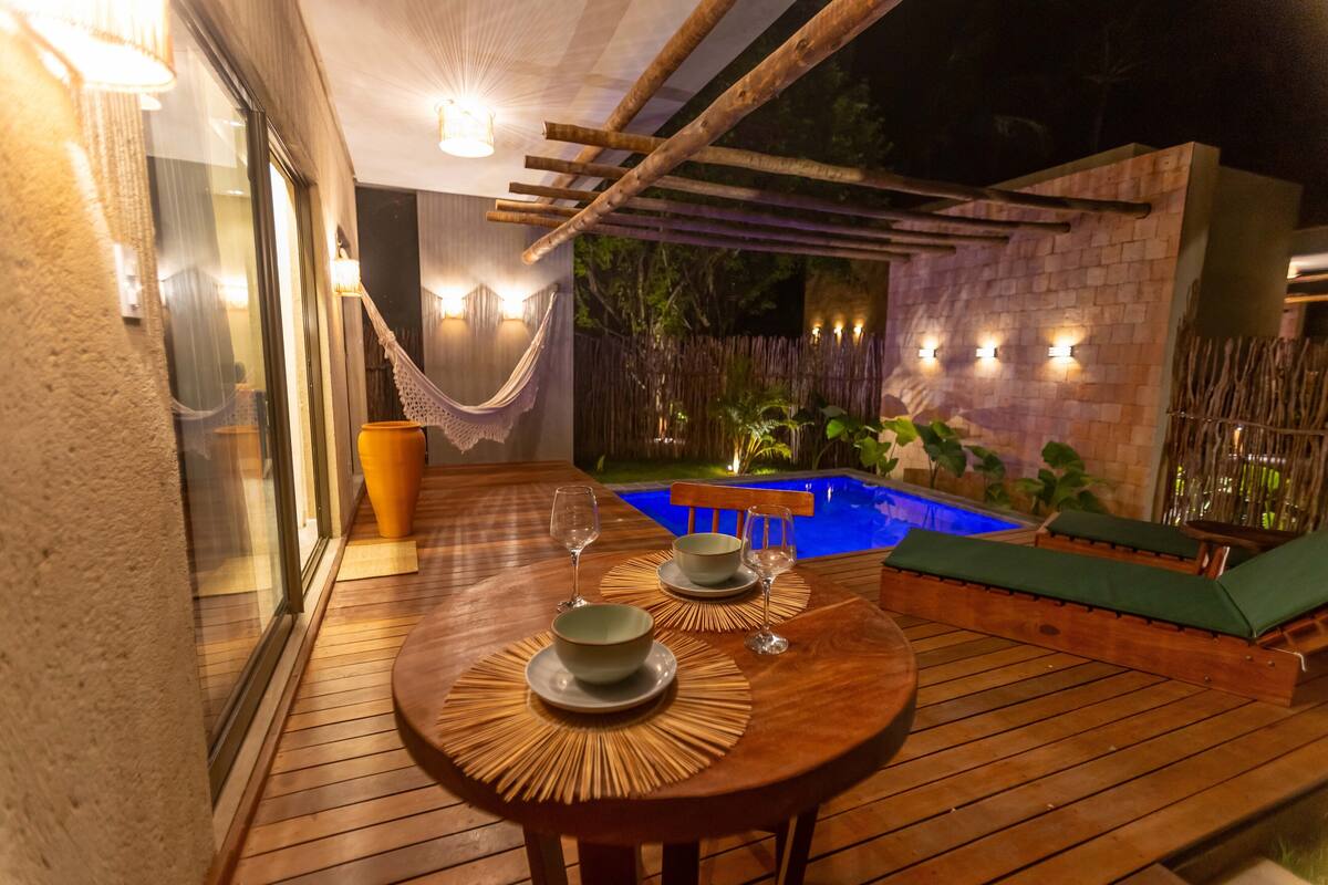 An outdoor dining area with a wooden table and chairs, a hammock, and a pool in the background.