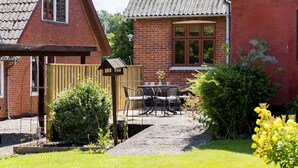 Une maison en briques avec une façade rouge, une clôture en bois, un jardin fleuri et une table avec des chaises à l’extérieur.