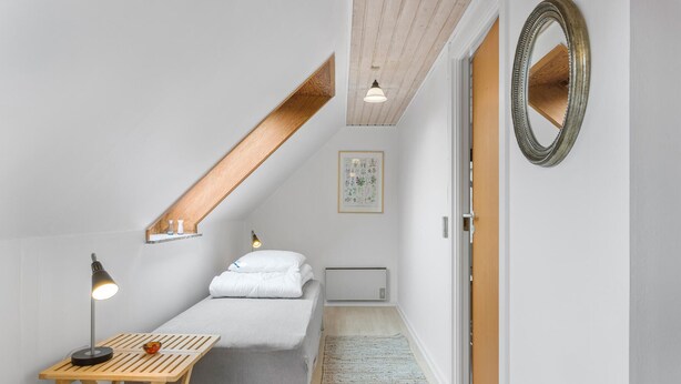 Une petite chambre bien éclairée, avec un plafond en pente, un bureau en bois, un lit et un miroir.