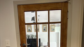 Une porte vitrée à cadre en bois donne sur un salon où se trouvent un piano, des canapés et des tableaux encadrés accrochés au mur.