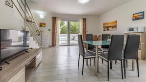 Un comedor moderno con una mesa y sillas de vidrio, un televisor sobre una encimera de madera y vistas a una piscina a través de grandes puertas de cristal.