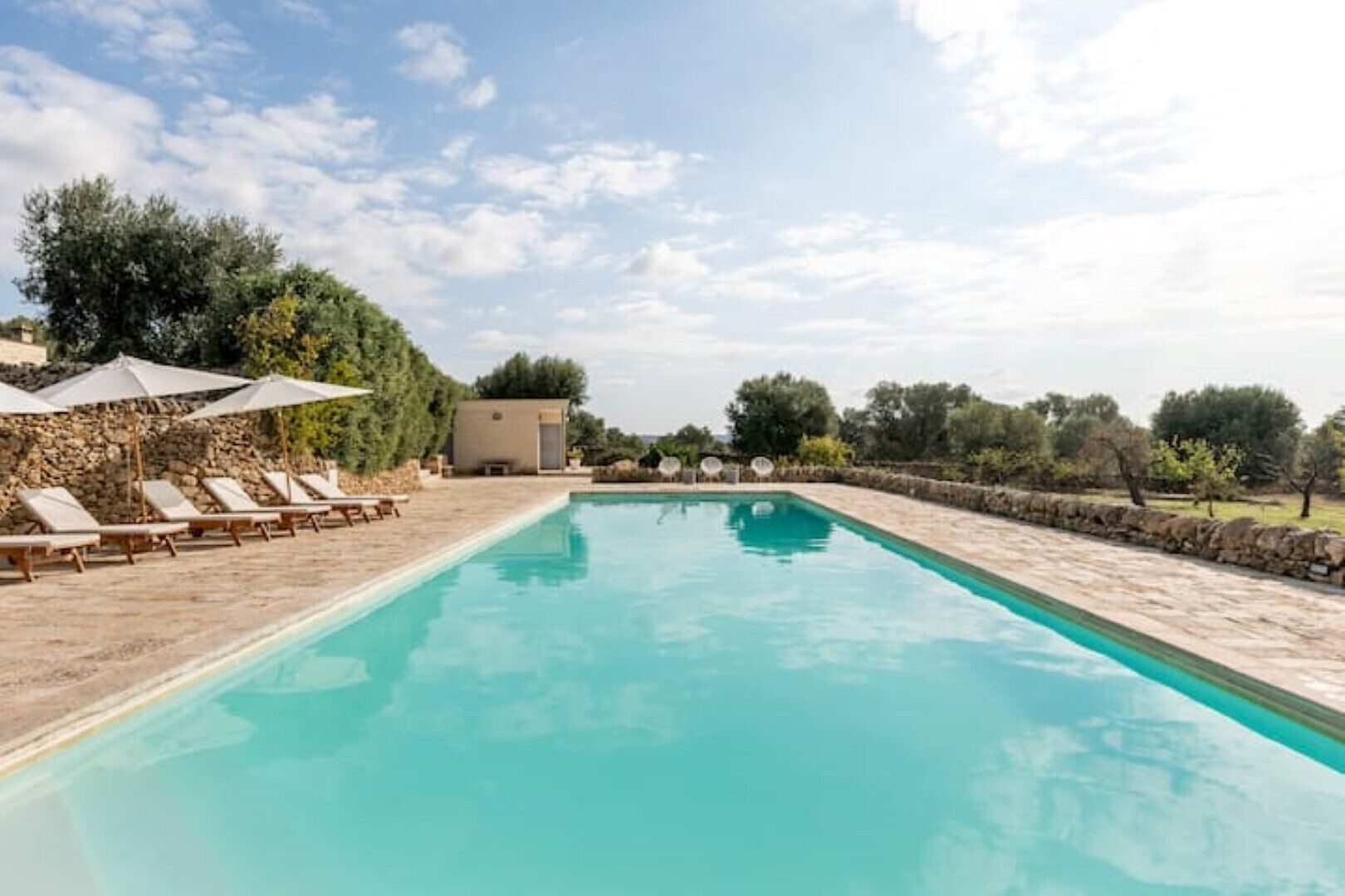 Une piscine rectangulaire à l’eau d’un bleu limpide, entourée de chaises longues et de parasols, avec en arrière-plan des arbres et un mur de pierre.