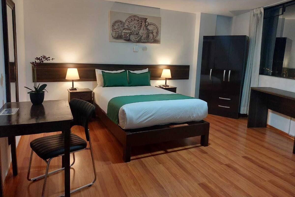 Une chambre d’hôtel comprenant un lit, deux tables de chevet avec des lampes, un bureau avec une chaise, une plante en pot, un grand miroir, une armoire et une œuvre d’art accrochée au mur.