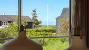 A view through a window with a white lamp in the foreground, a grassy area with bushes and a flagpole in the background, and a building with a dark roof. - 3 bedroom cozy home in Lundby (Lundby)