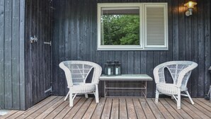 Puinen terassi, jossa on kaksi valkoista rottinkituolia ja pieni pöytä, ikkuna, jossa on sälekaihtimet, ja seinälle ripustettu lyhty.