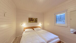 Una camera da letto con soffitto in listelli di legno, un letto con lenzuola bianche, un quadro appeso al muro e una finestra con vista sugli alberi.