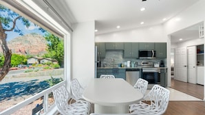 Une cuisine moderne avec des meubles blancs et un grand îlot central, un coin repas avec des chaises blanches, et une vue sur un paysage montagneux grâce à de grandes fenêtres.