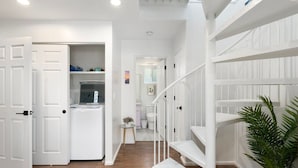 Un intérieur aux murs blancs, avec un escalier, une porte donnant sur une salle de bain et une petite table sur laquelle est posée une plante.