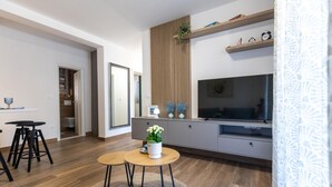 Een moderne woonkamer met een televisie, houten details, een eethoek met krukken, en een keuken op de achtergrond.