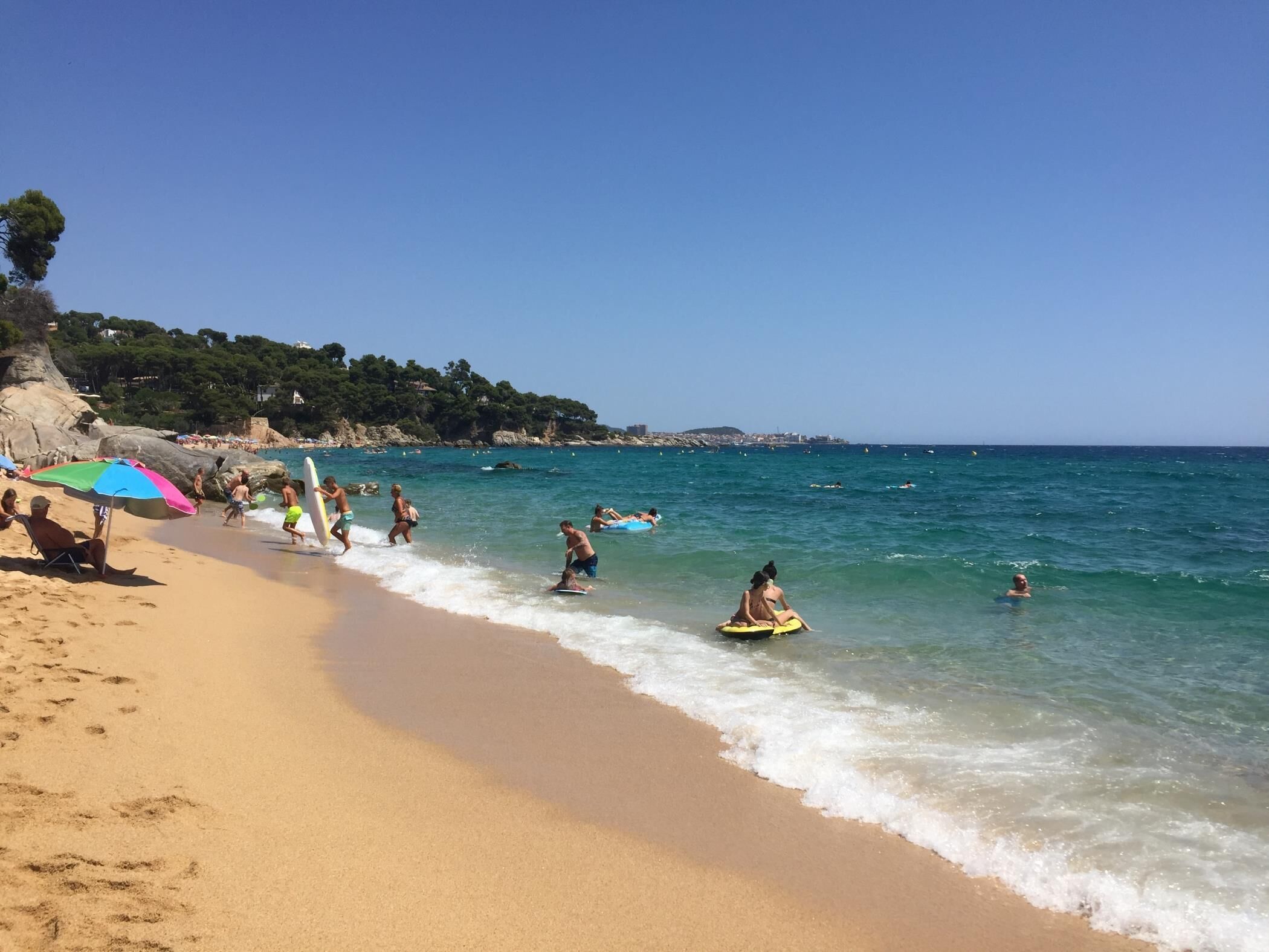 Ein Strand mit badenden und spazierenden Personen, ein klarer Himmel und eine felsige Küste im Hintergrund.