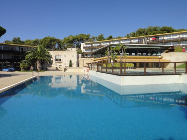A swimming pool with a clear blue water surface, surrounded by a building with multiple balconies and a covered area with seating.