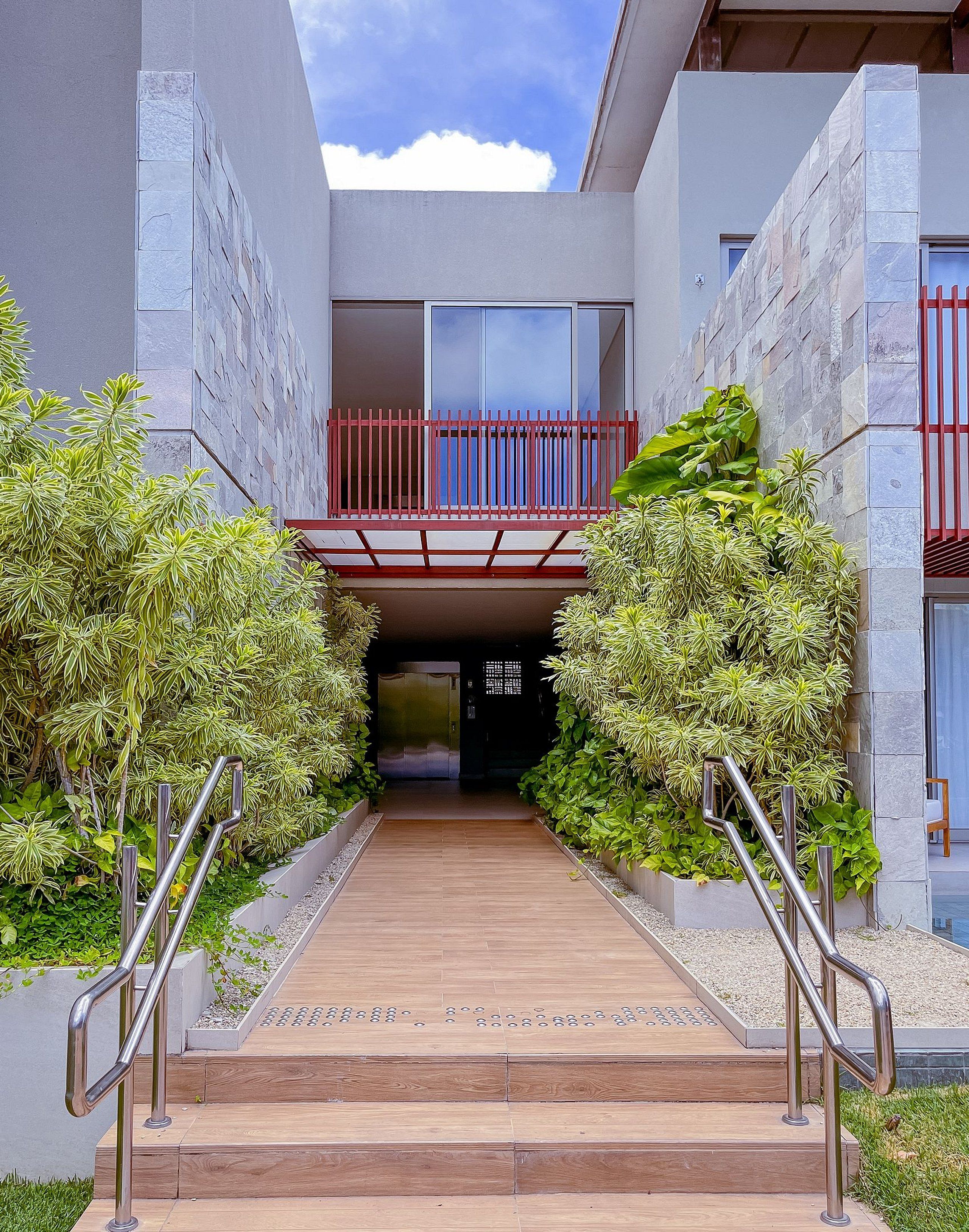 Una entrada de edificio moderna con un sendero de madera, barandas de metal y una barandilla de balcón roja.