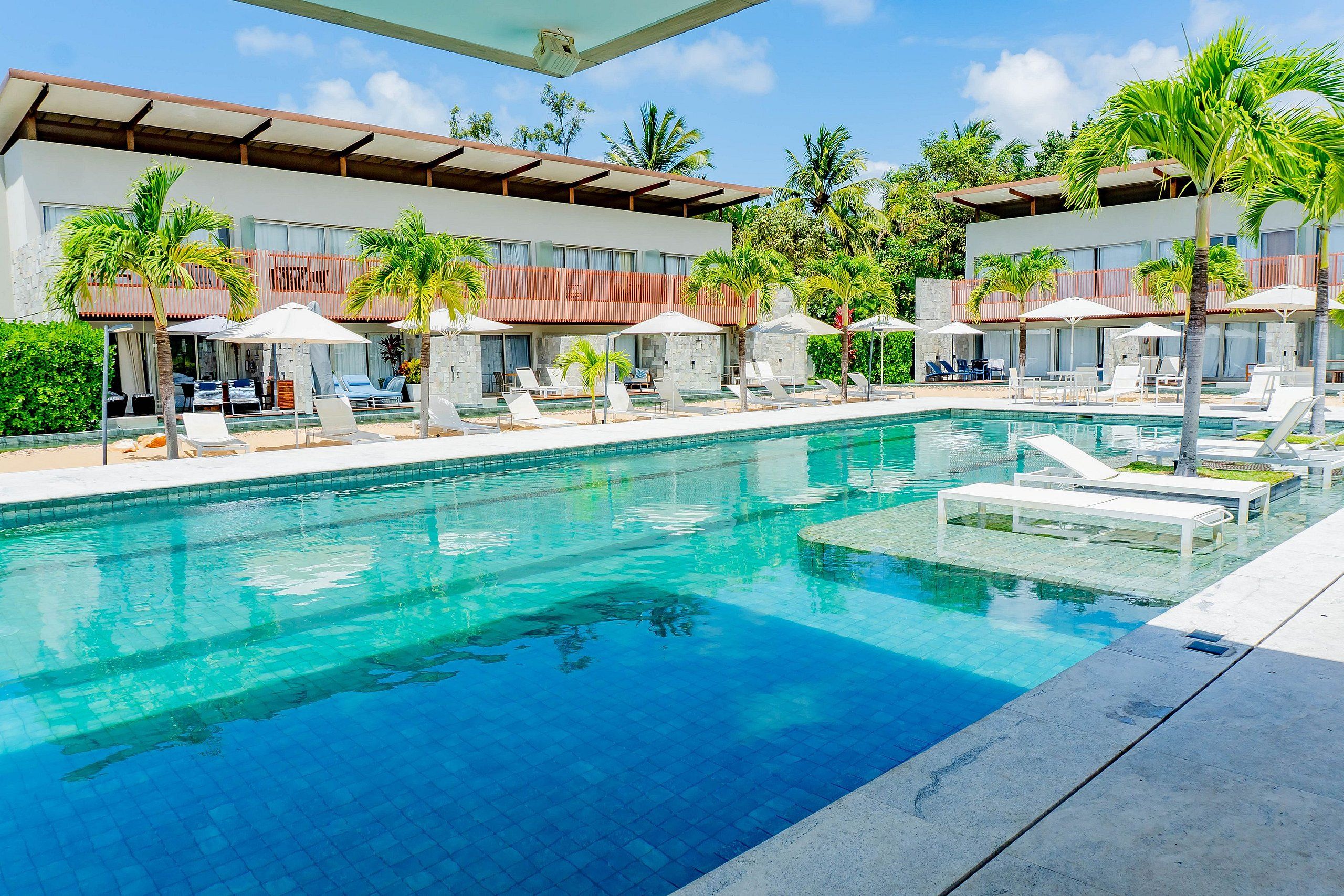 A swimming pool with lounge chairs and umbrellas, surrounded by palm trees and a building with a red roof.