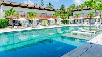 A swimming pool with lounge chairs and umbrellas, surrounded by palm trees and a building with a red roof.