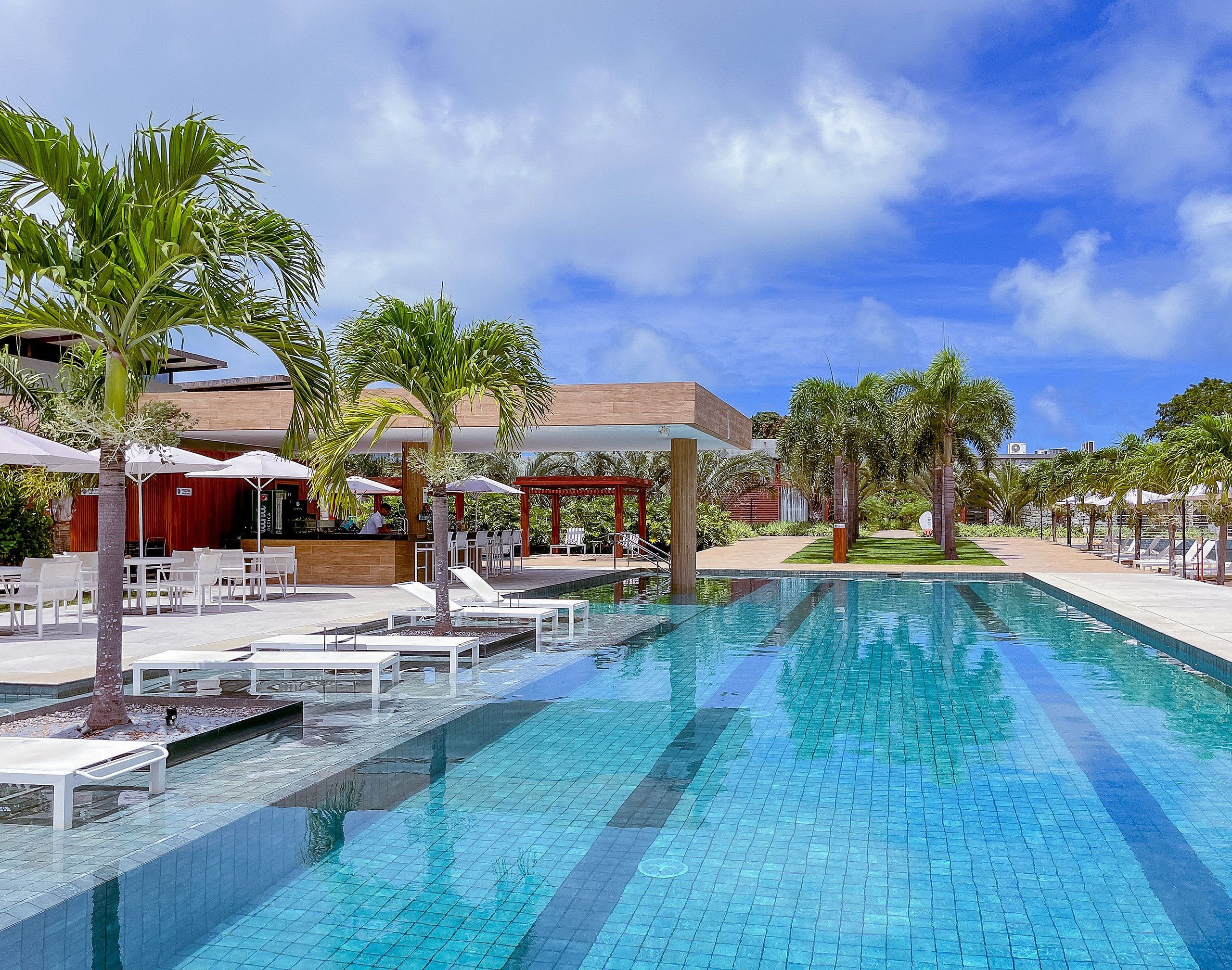 Piscina com espreguiçadeiras e guarda-sóis, cercada por palmeiras e um edifício com fachada vermelha.