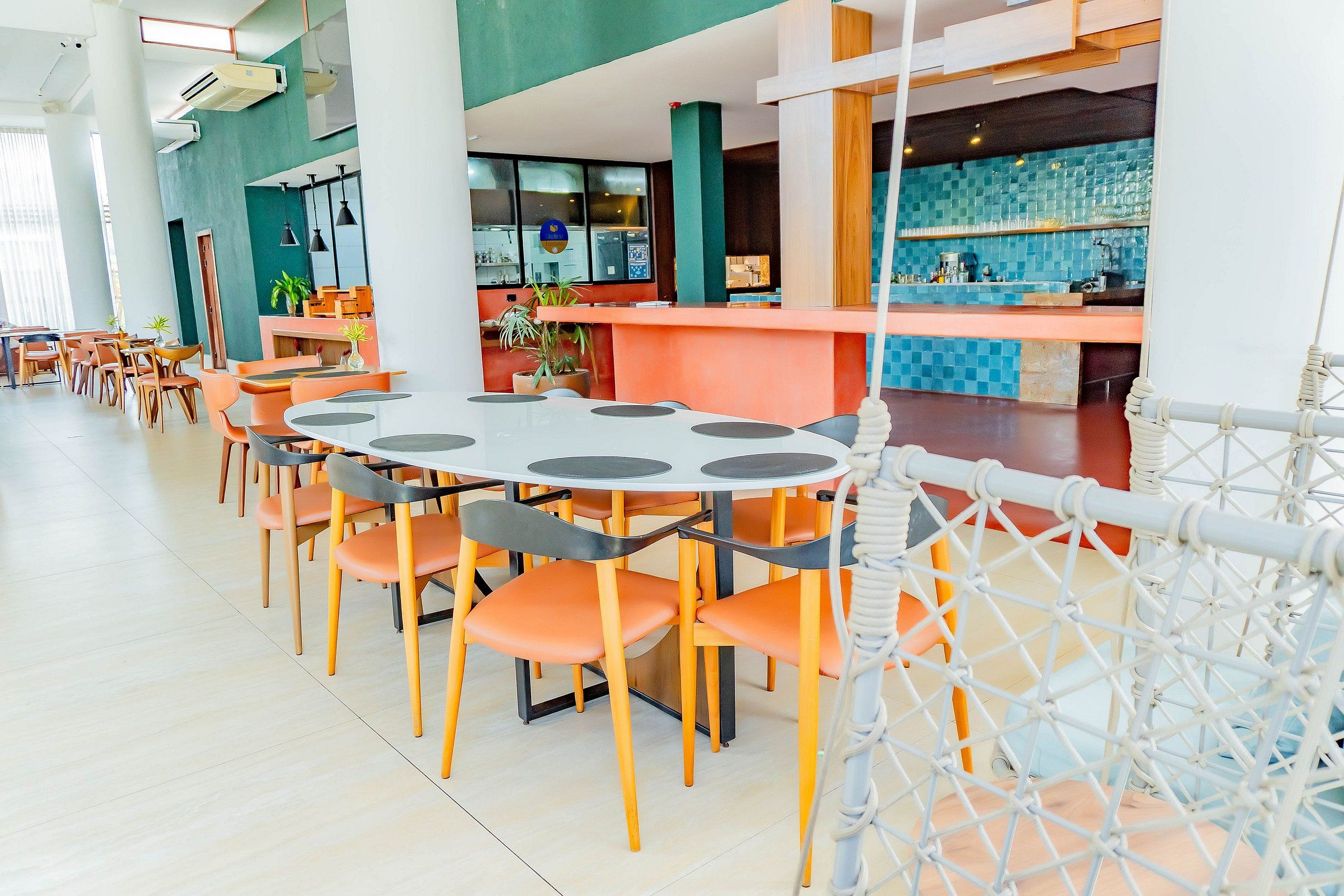 Un restaurante moderno con mesas redondas y sillas naranjas, una zona de bar con azulejos azules y una barrera de cuerda blanca.