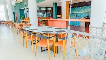 Un restaurante moderno con mesas redondas y sillas naranjas, una zona de bar con azulejos azules y una barrera de cuerda blanca.