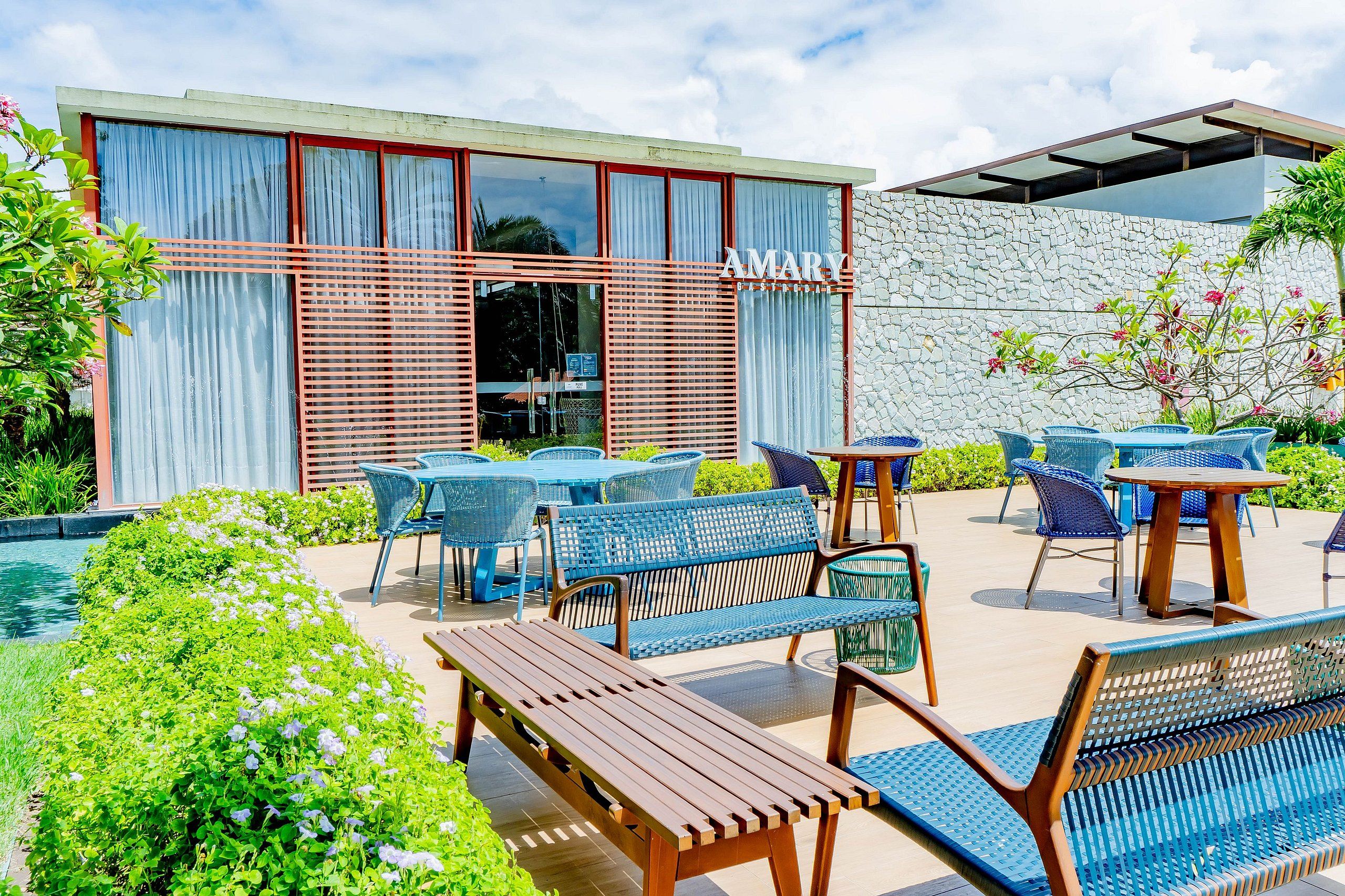 An outdoor seating area with tables and chairs, a pool, and a building with a stone wall and a sign that reads 'AMARY GASTRO CLUB'.