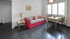 A room with a red sofa, a small table with a lamp, a dining table with chairs, and a window with blinds. - La casa del Cartografo (Genova)