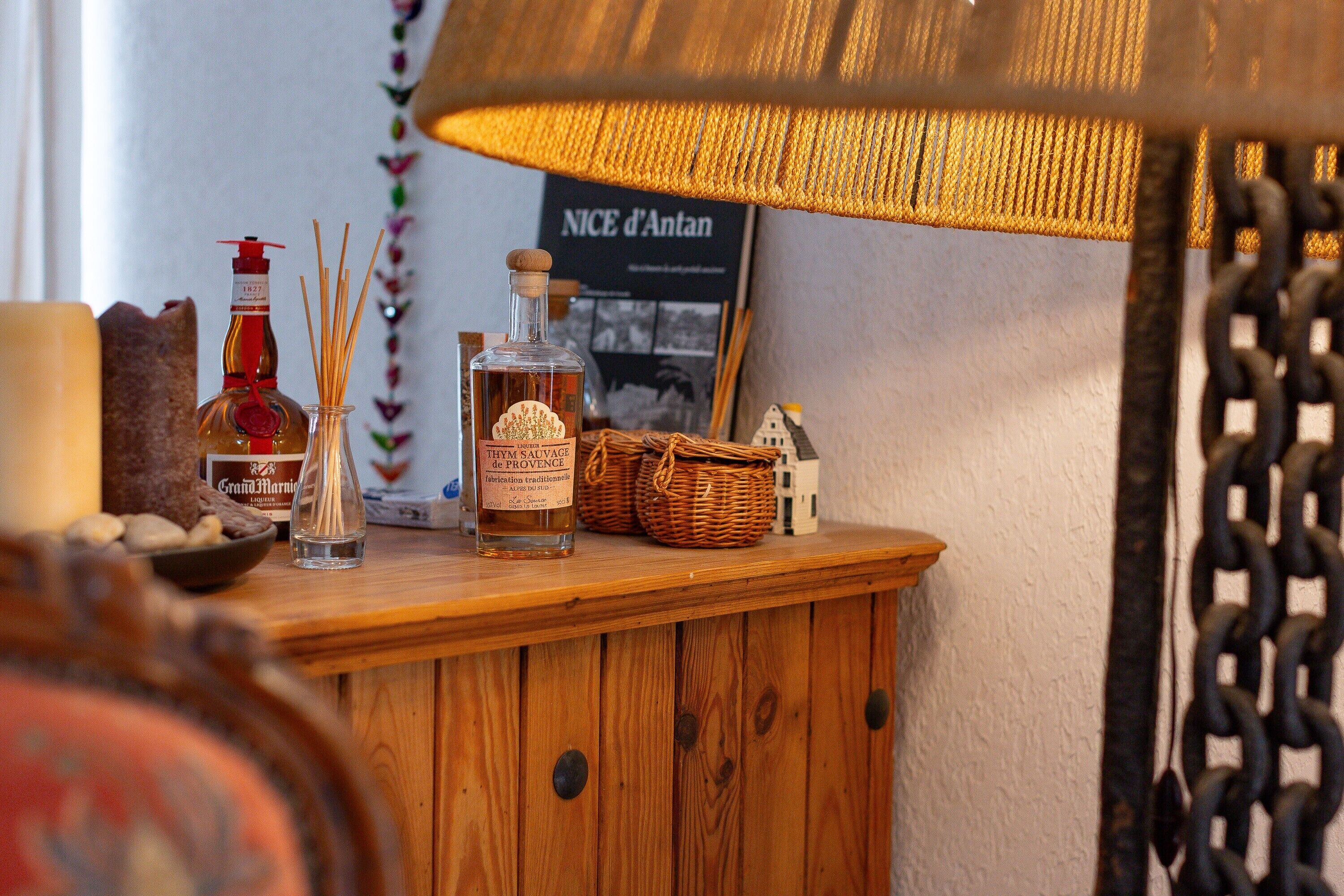 Un meuble en bois surmonté d’une lampe, une bouteille de thym de Provence, un petit panier et une maquette de maison miniature.