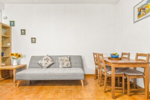 A living room with a grey sofa, a wooden dining table with chairs, a small side table, and a wall-mounted picture frame.