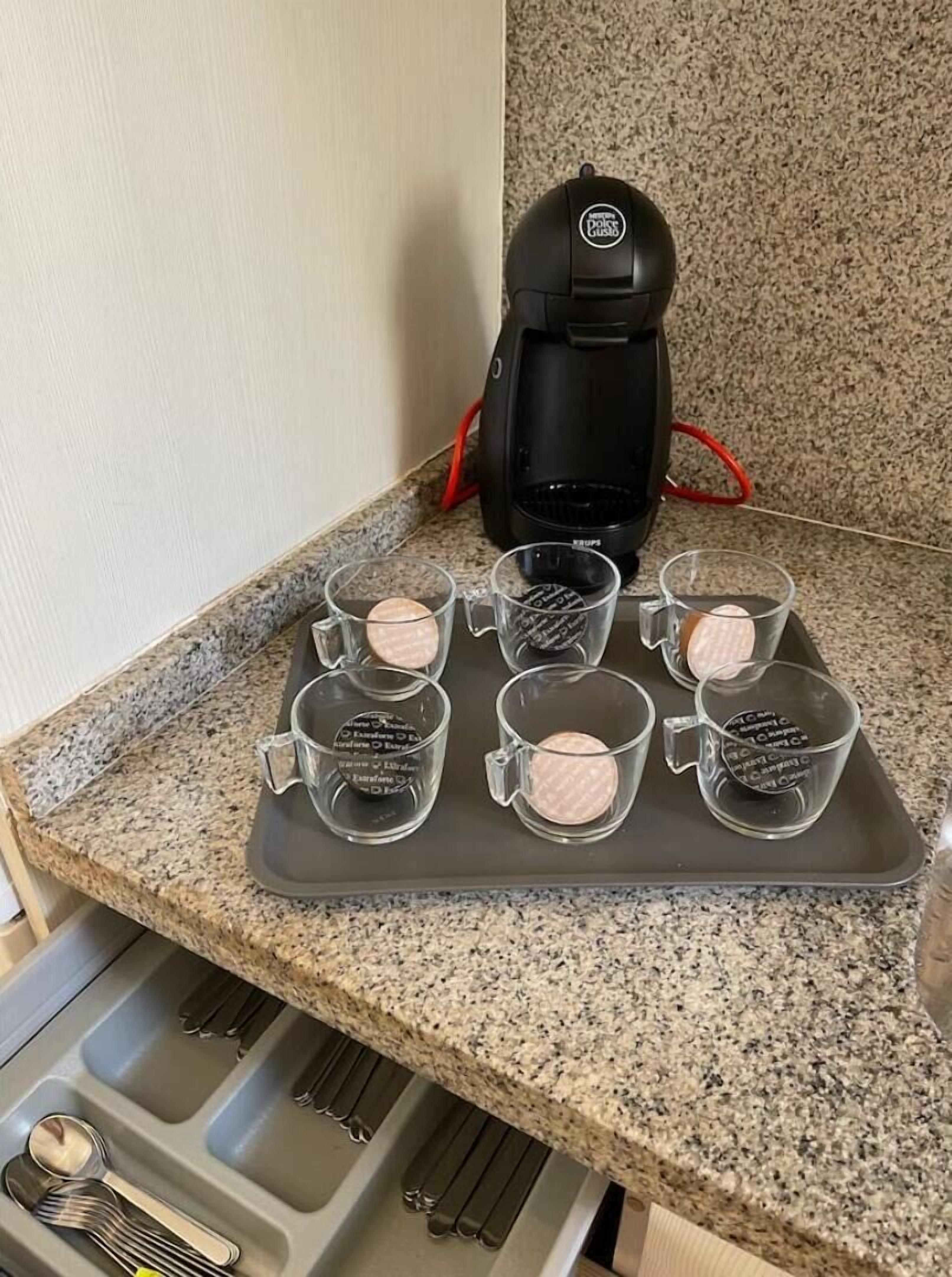 A kitchen counter with a coffee maker, a tray with six glass mugs, and a set of utensils.