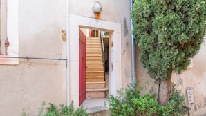 Un escalier étroit menant à une porte ornée d’un volet rouge, un petit arbre et un luminaire fixé au mur.