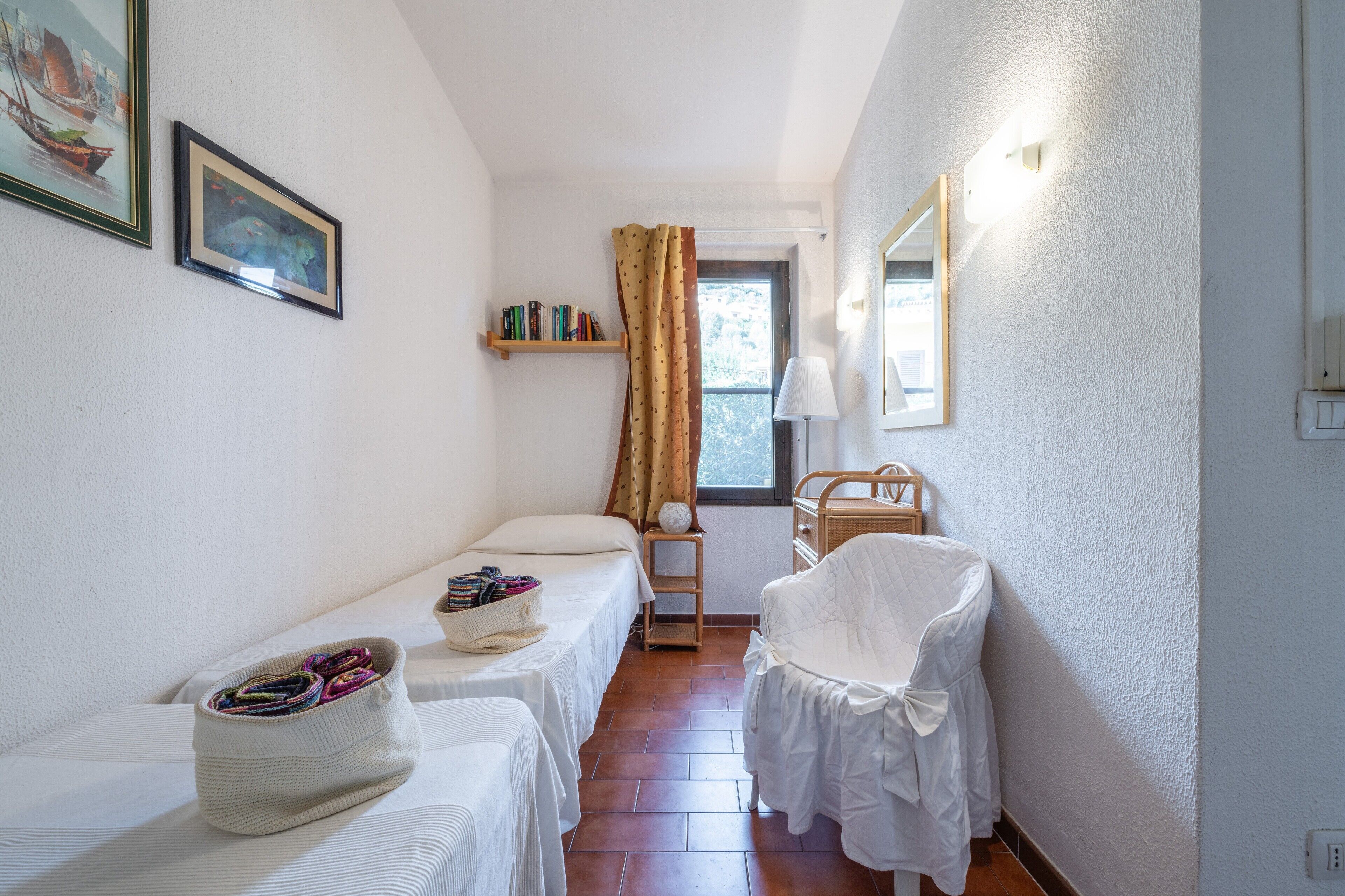 A narrow room with two beds, a chair, a shelf with books, a window with curtains, and a lamp.