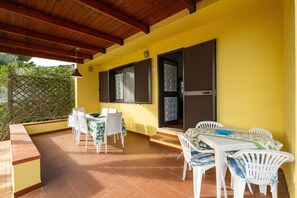 Une terrasse couverte avec un mur jaune, deux fenêtres avec volets, une table avec des chaises et une vue sur la verdure.
