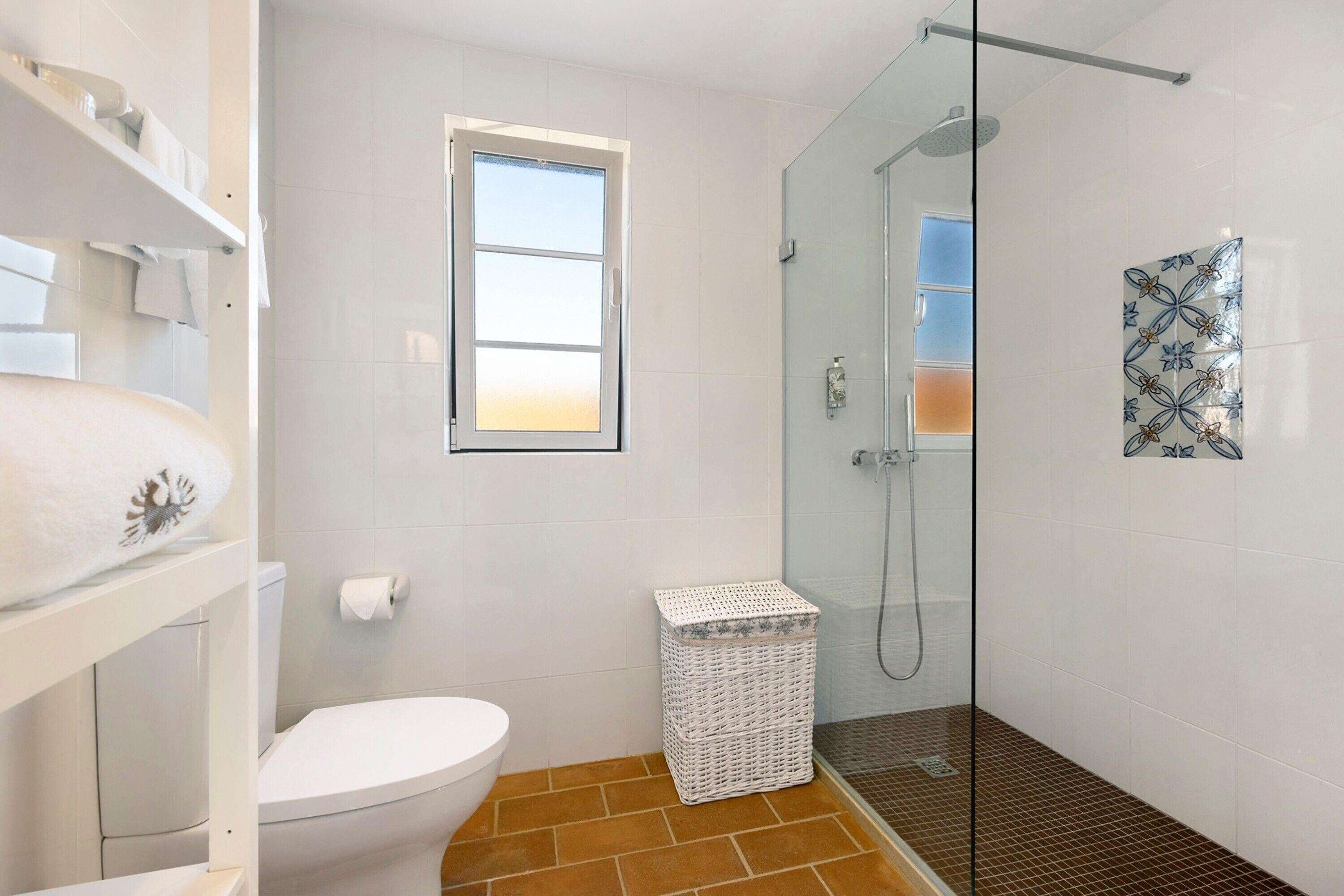 A bathroom with a toilet, a glass shower enclosure, a small window, and a white wicker basket.