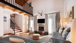 Un salon avec un escalier, une cheminée, un téléviseur et une tête de cerf fixée au plafond.