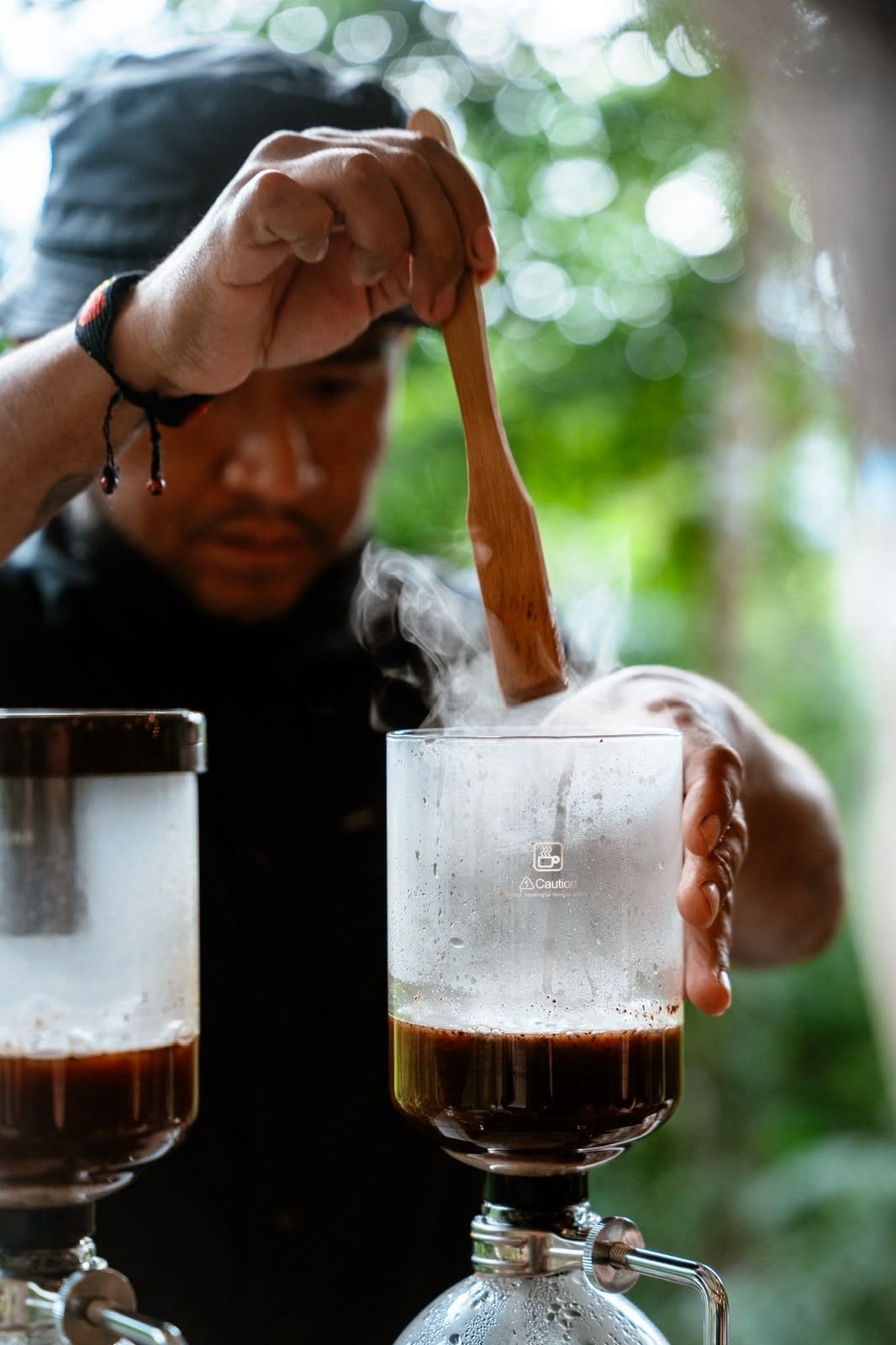 A person pouring a dark liquid into a glass with a wooden spoon, with steam rising from the glass.