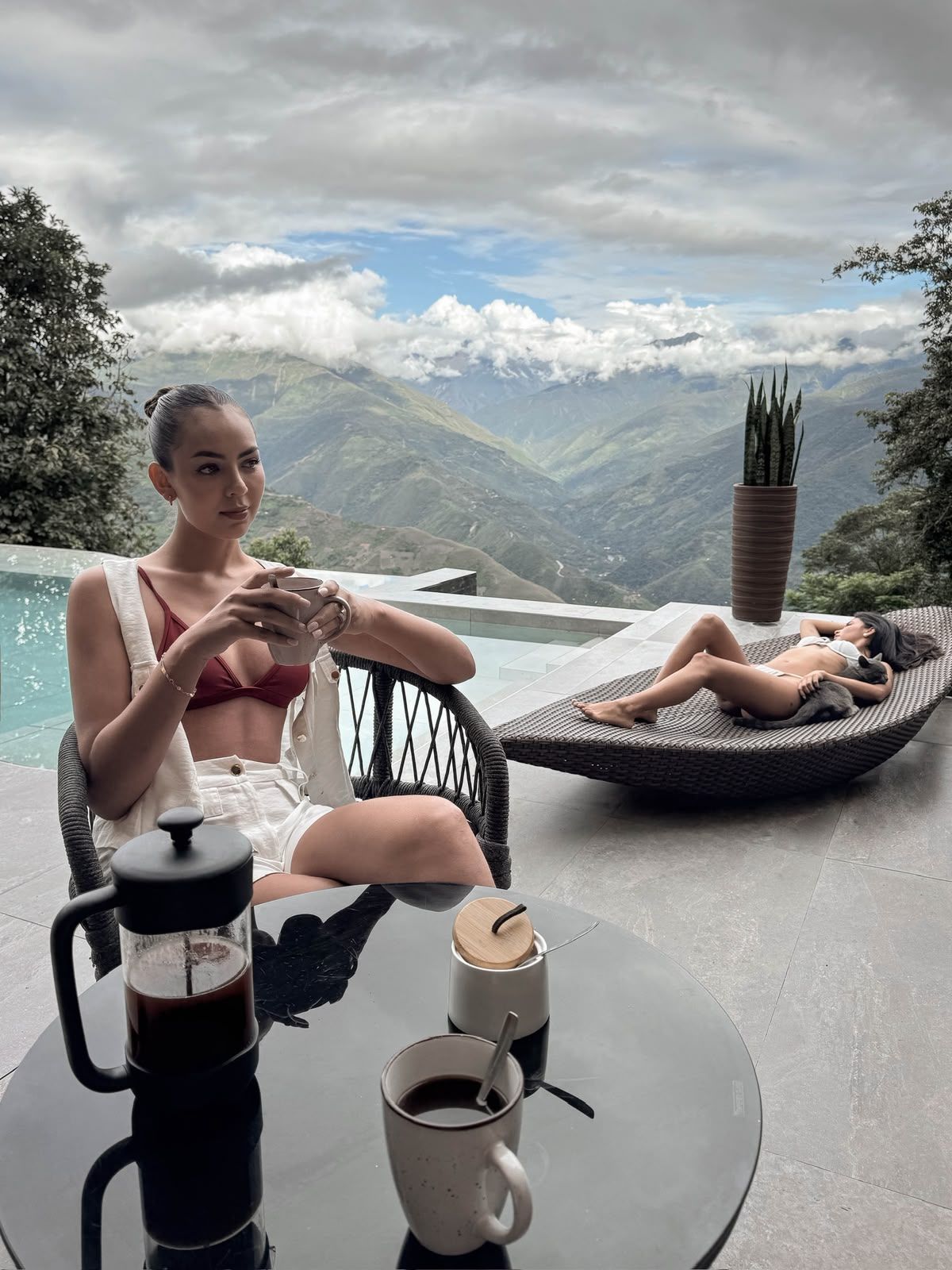 A person sitting at a table with a coffee pot and cup, another person lying on a lounge chair with a plant beside them, and a view of mountains and clouds in the background.