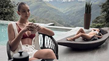A person sitting at a table with a coffee pot and cup, another person lying on a lounge chair with a plant beside them, and a view of mountains and clouds in the background.