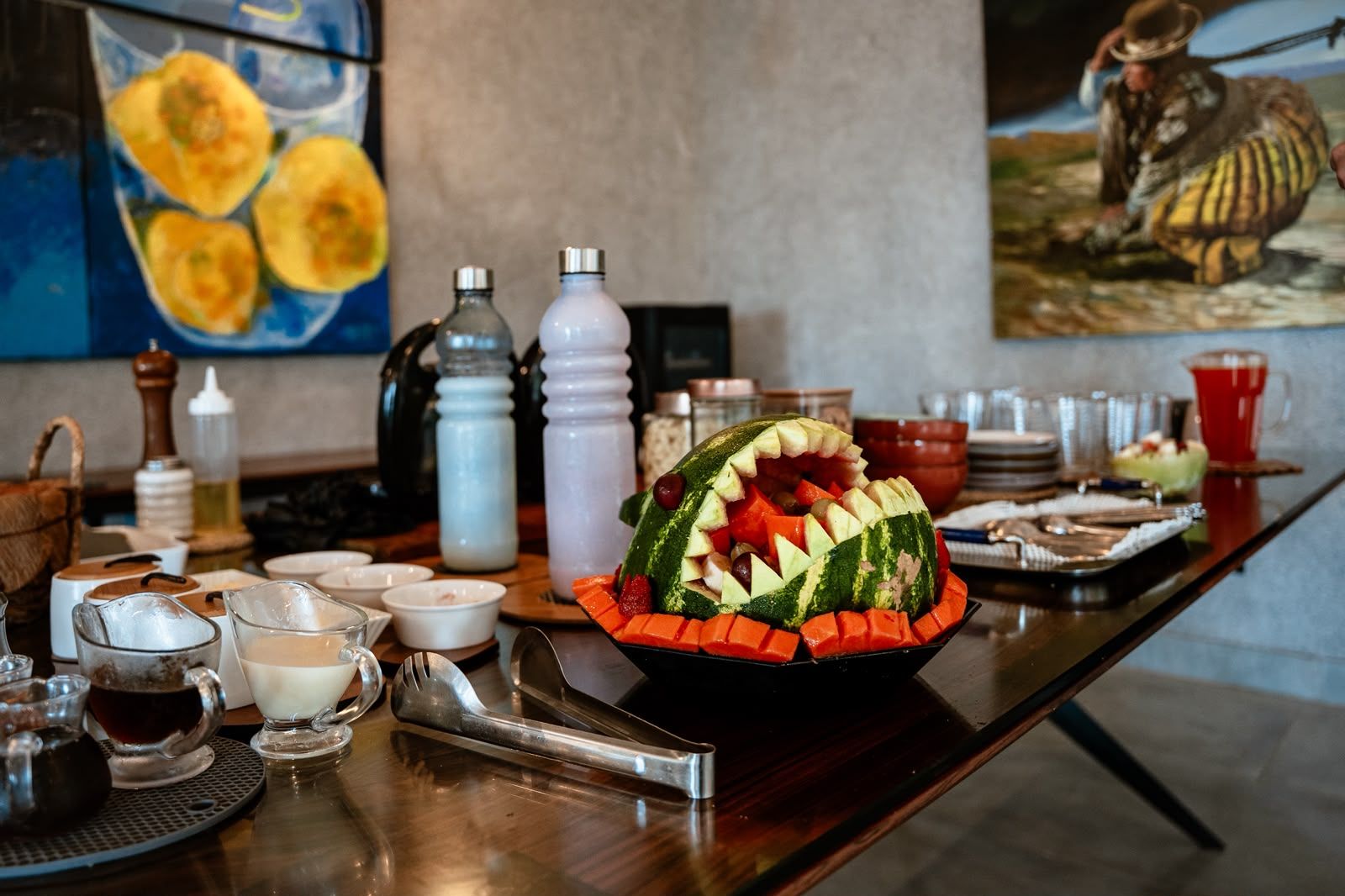 A table with a watermelon carved into a shark, surrounded by various food items and condiments.