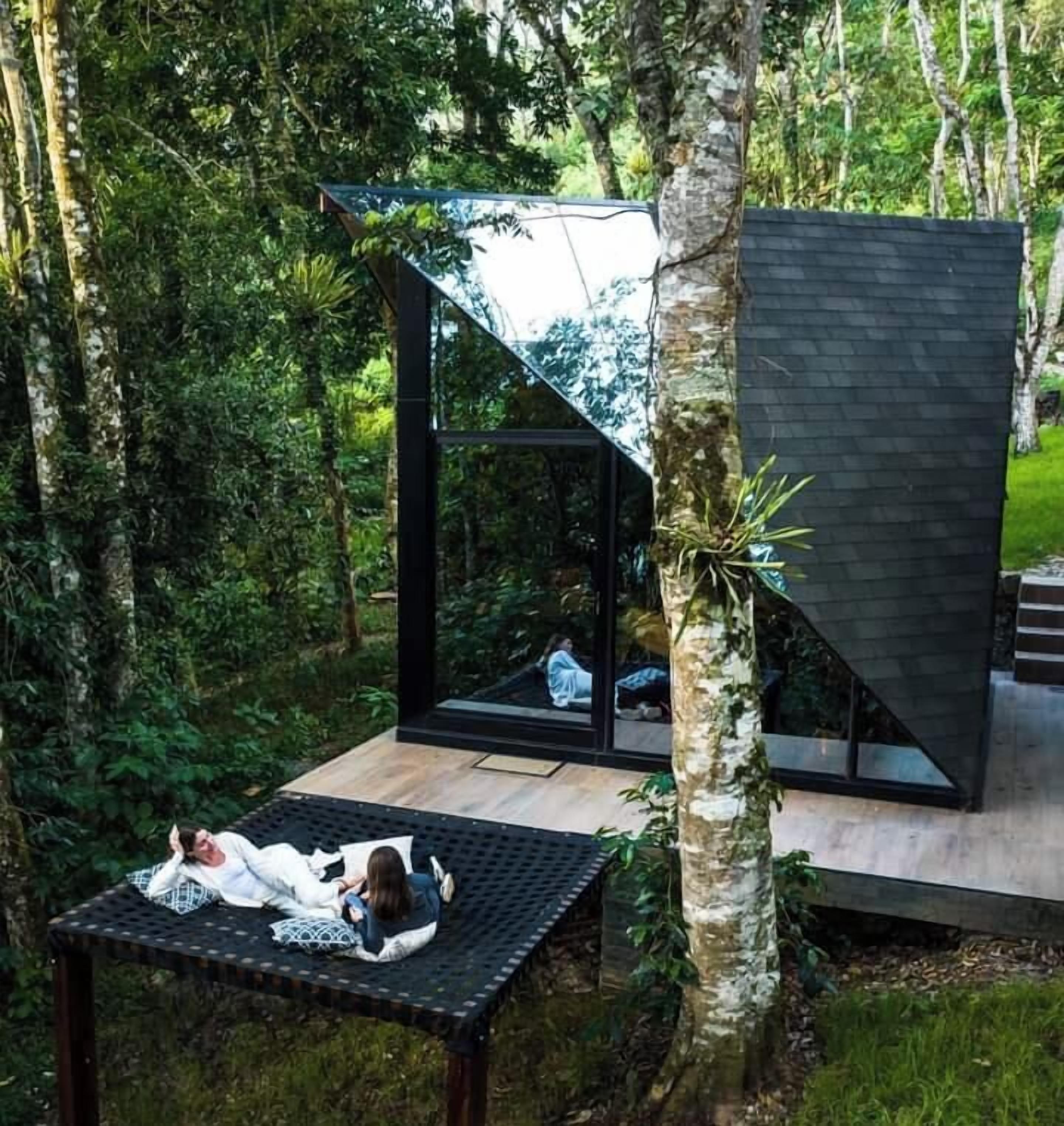 Een moderne boomhut met een glazen omheining, een houten terras en een hangmat.