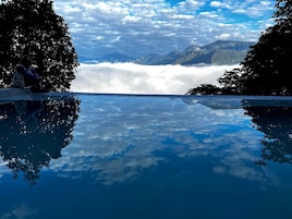 Vista despejada de una cordillera con nubes abajo, un estanque que refleja el paisaje y una persona sentada en un saliente.