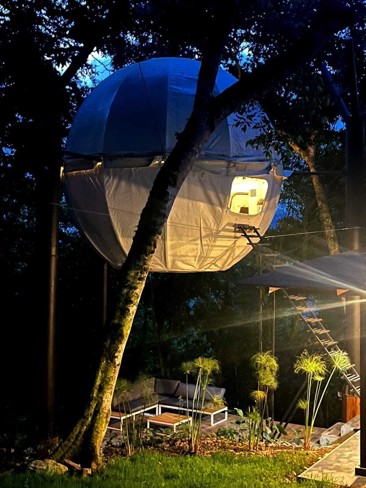 A spherical structure with a ladder leading to a door, surrounded by trees and outdoor furniture.