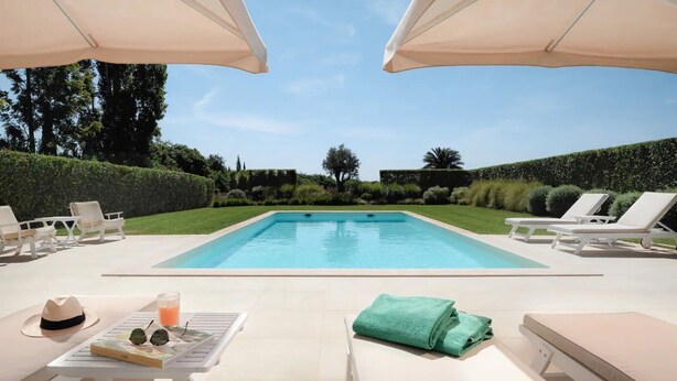 Una piscina con sillas de descanso y sombrillas, rodeada de follaje.