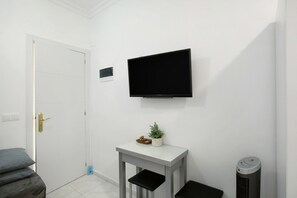 Une pièce blanche avec une porte fermée, un téléviseur fixé au mur, une petite table avec une plante et un radiateur portatif.