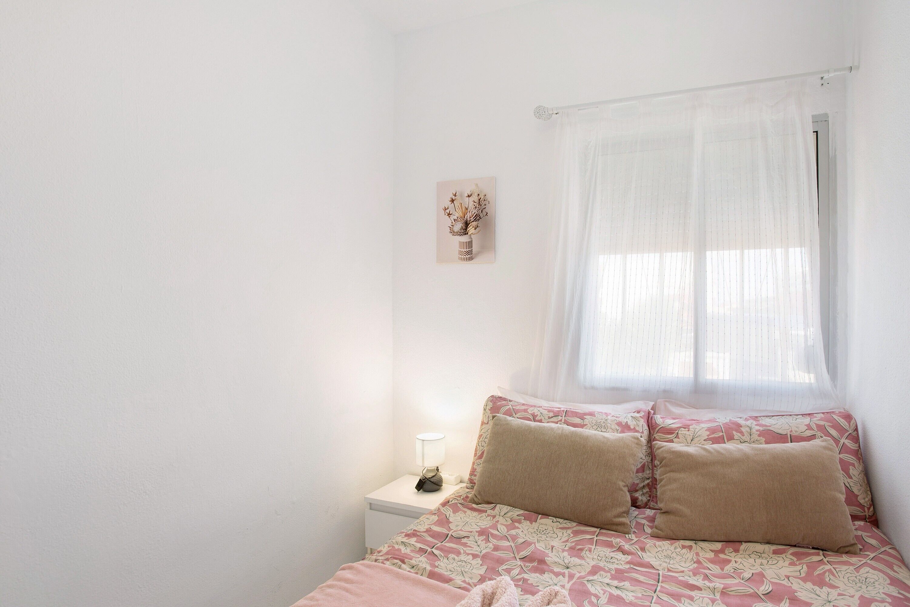 Une chambre à coucher avec un lit, une table de chevet avec une lampe, une fenêtre avec des rideaux légers et un tableau accroché au mur.