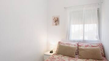 Une chambre à coucher avec un lit, une table de chevet avec une lampe, une fenêtre avec des rideaux légers et un tableau accroché au mur.