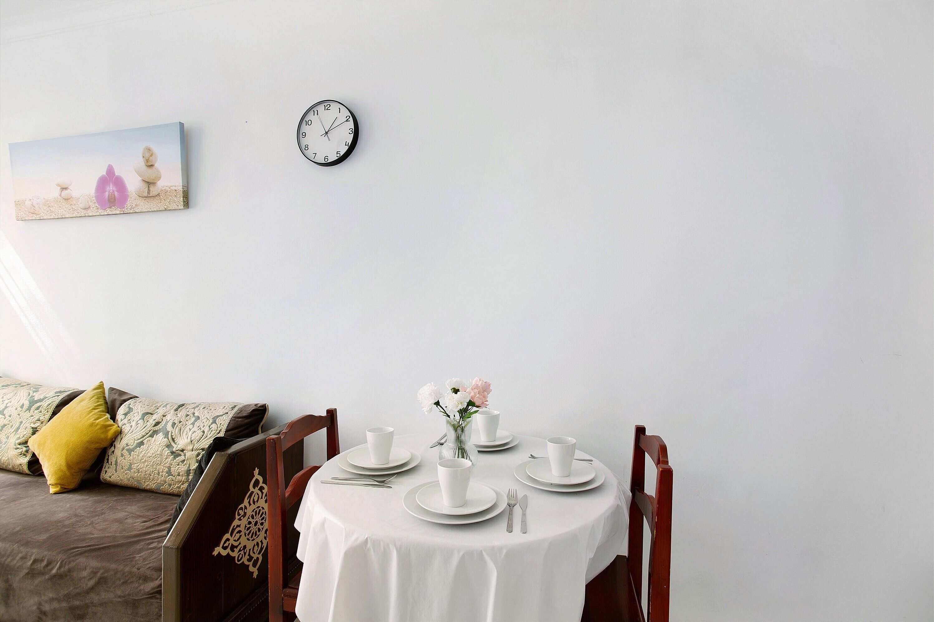 A dining area with a round table set for four, a clock on the wall, and a bed with pillows in the background.